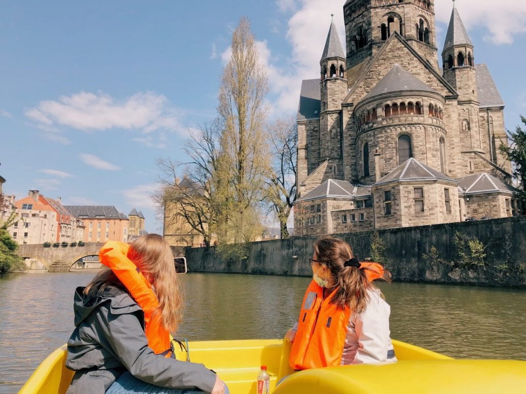An image of Kaitlyn and another friend sitting in a bright yellow paddle boat on the river. Their backs are to the camera and they are looking to the right at the Temple Neuf from the water.