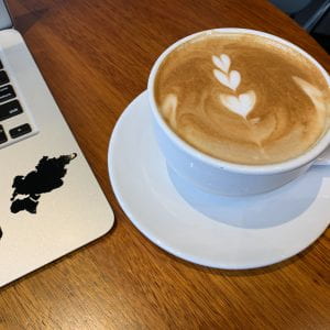Wooden table with a light brown latte in a white mug on a white plate. The latte art is 3 white hearts in a vertical line. To the left of the mug is the corner of a silver laptop keyboard with a half peeled off sticker of Eurasia and Africa.