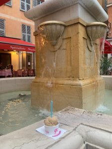 Image Description: White cup with a pink logo full of light brown gelato and a teal spoon, on a stone ledge in front of a running fountain. In the background is an orange building with a red cloth overhang with white text: "Spécialité Niçoise."