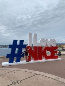 ID: A woman with a maroon top and black shorts, spreading her arms in front of a large blue, white, and red sign that reads: "#ILoveNice." Behind the sign, the sea and coastline is visible, and the sky is overcast.