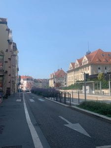 Road lined with pale colored buildings with red brick roofs. In the median of the road are purple flowers. The sky is a pale blue with no clouds.
